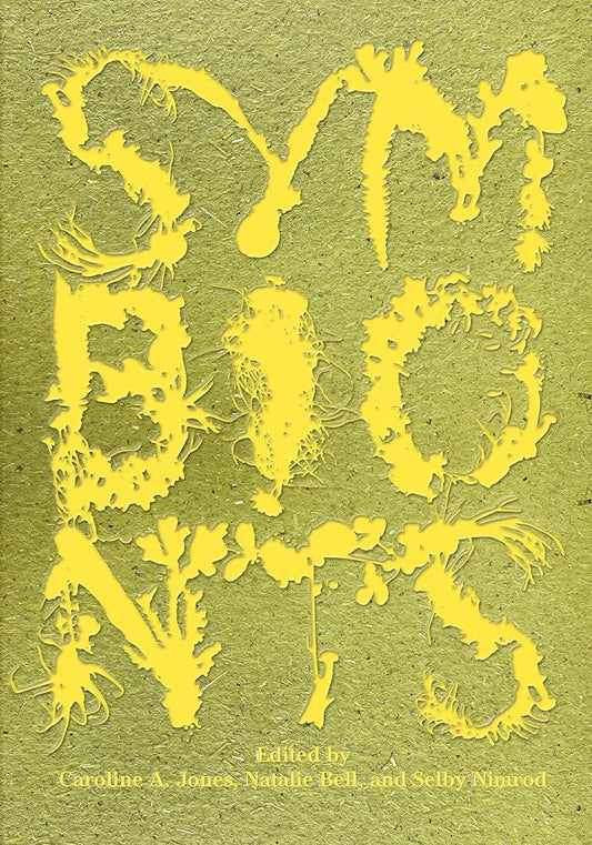 The image features a close-up view of a book cover with the title "Symbionts: Contemporary Artists and the Biosphere." The cover displays a vibrant, abstract design in yellow and green tones, with the word "SYMBIONT"S" prominently featured in a stylized font.
